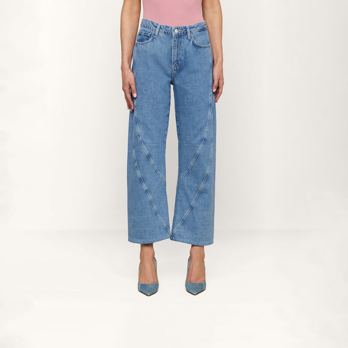 A person wearing the Frasier Rodeo Jean—slouchy-fit, wide-leg blue jeans with curved seam details—paired with a pink top and blue pointed-toe heels, stands against a plain white background.
