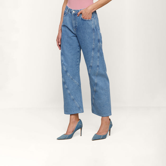 A person wearing the Frasier Rodeo Jean, a light blue high-rise denim wide-leg jean with decorative seams, along with a pink top and blue high heels, stands against a plain white background.