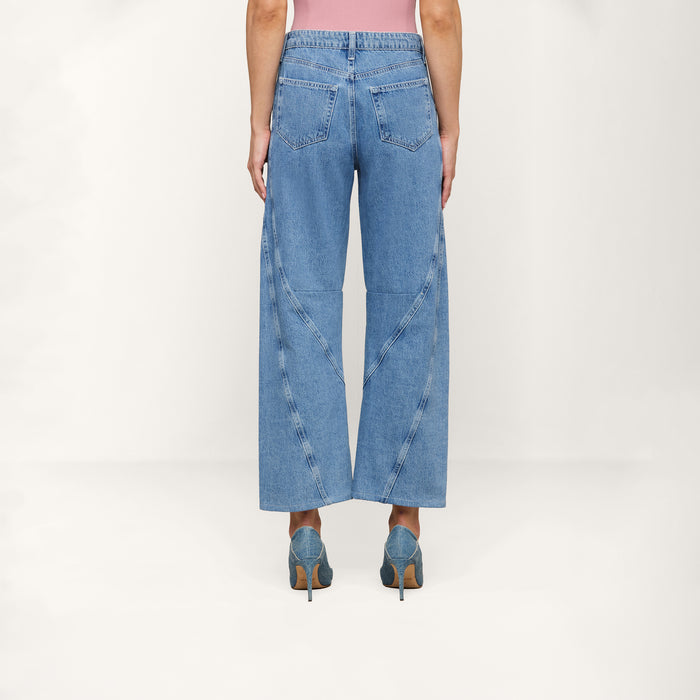 Person standing with their back facing the camera, wearing a pink top, Frasier Rodeo Jean—a slouchy-fit wide-leg blue jean with unique seam details—and blue high-heeled shoes on a plain light background.