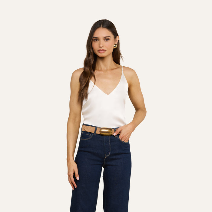 A woman with long brown hair wears the Gabriella Silk Camisole—a white sleeveless V-neck top—paired with dark blue jeans, a brown belt, and gold hoop earrings, standing against a plain light background.