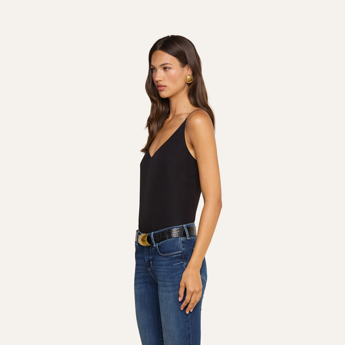 A woman in profile stands against a plain light background, wearing the Gabriella Silk Camisole in black paired with blue jeans. She accessorizes with large round earrings and a black belt featuring a gold buckle, her hair worn down.