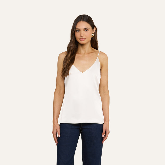 A woman stands facing forward against a plain light background, wearing the Gabriella Silk Camisole in ivory—a V-neck design—paired with dark blue pants. Her long brown hair and gold hoop earrings complete the look.