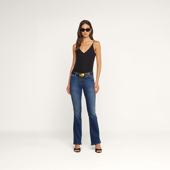 A woman stands confidently against a plain white background, wearing sunglasses, blue flared jeans, black heels, and the Gabriella Silk Camisole.