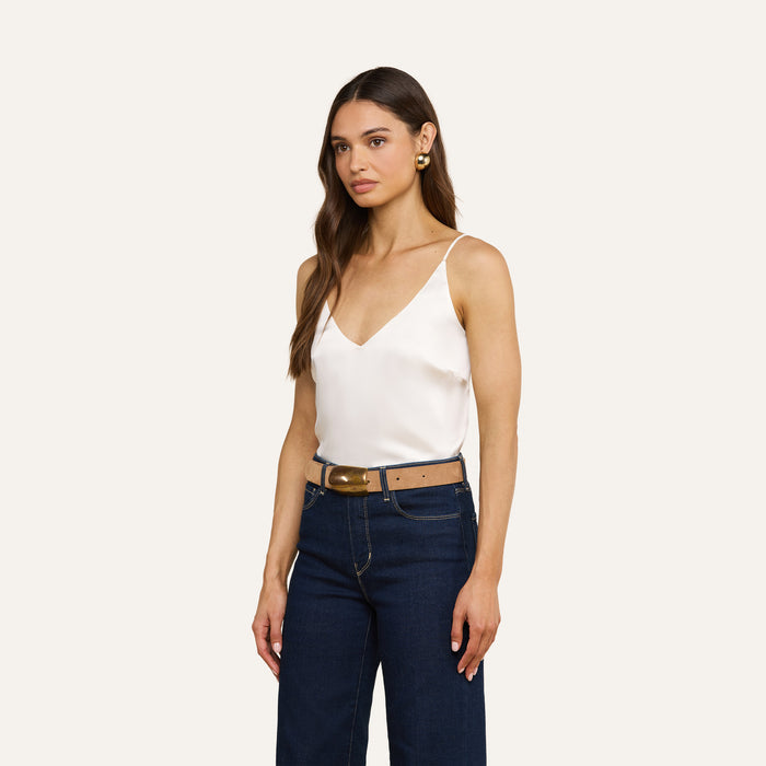 Woman with long brown hair wearing the Gabriella Silk Camisole in ivory, paired with dark blue jeans, a brown belt, and gold earrings, standing against a plain light background.