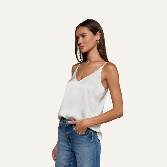 A woman with long brown hair wears the Gabriella Silk Camisole in ivory, paired with blue jeans, standing with one hand in her pocket against a plain light background.