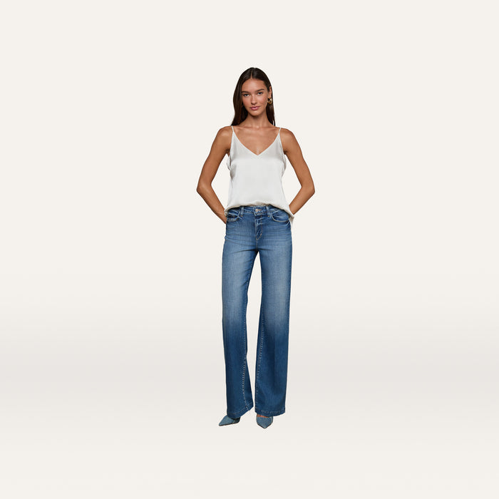 A woman stands against a plain white background with her hands in the pockets of her blue flared jeans, wearing the Gabriella Silk Camisole in ivory.