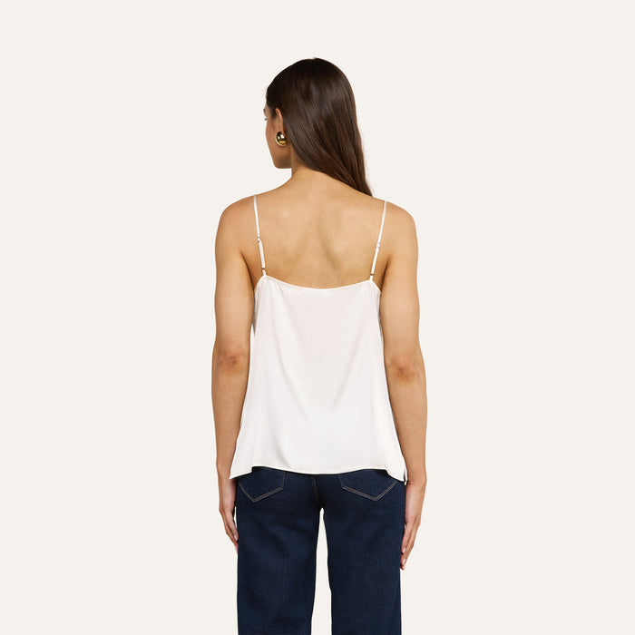 A woman stands facing away, wearing the Gabriella Silk Camisole in white paired with dark blue jeans. Her long brown hair is down, and she accessorizes with gold hoop earrings. The background is plain and light-colored.