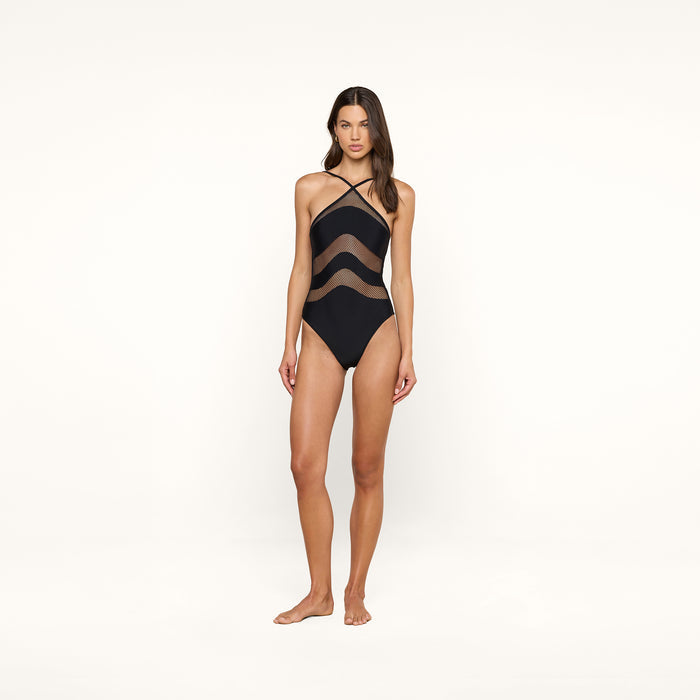 A woman stands barefoot against a plain white background, wearing the Gabrielle One-Piece Swimsuit featuring black fabric with mesh insets, sheer wavy panels, and crisscross straps.