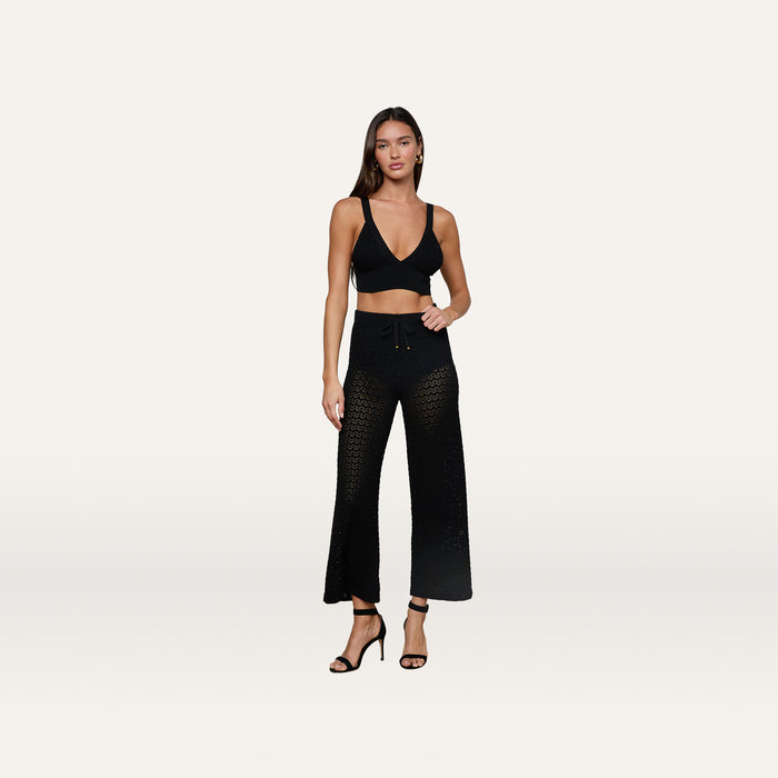 A woman stands against a plain white background wearing a black crop top, the Gaelle Pointelle Knit Pant in black featuring a sheer crochet knit and drawstring waist, and black high-heeled sandals.