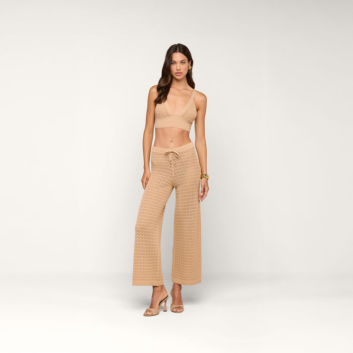 A woman stands in a neutral studio wearing a tan bralette top, high-waisted Gaelle Pointelle Knit Pants, and heeled sandals. She has long hair and a relaxed, confident pose—perfect for vacation vibes.
