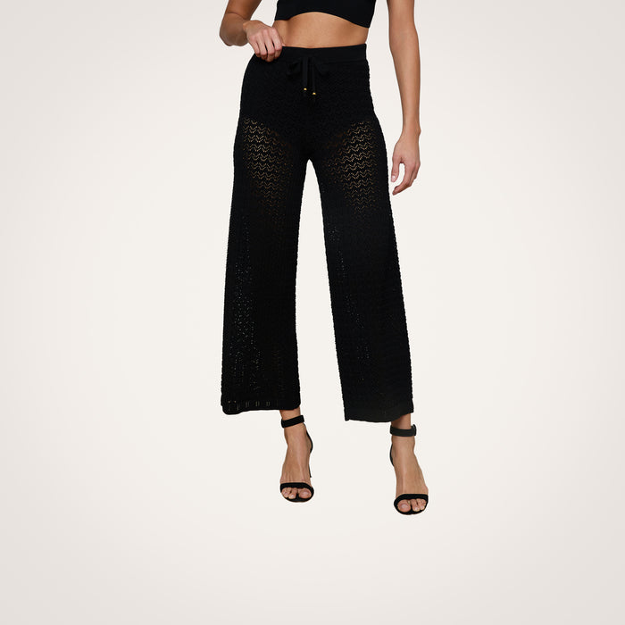 A woman wears the Gaelle Pointelle Knit Pant—a black, high-waisted, wide-leg knit pant with a drawstring waist—paired with a black crop top and black high-heeled sandals. Her face is not visible.