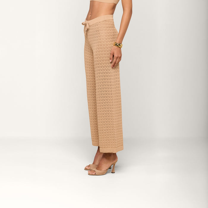 A woman models the Gaelle Pointelle Knit Pant, showcasing its beige wide-leg pointelle knit design with a tie waist, styled with heeled sandals and a bracelet against a plain white background. This lightweight piece is ideal for vacation.