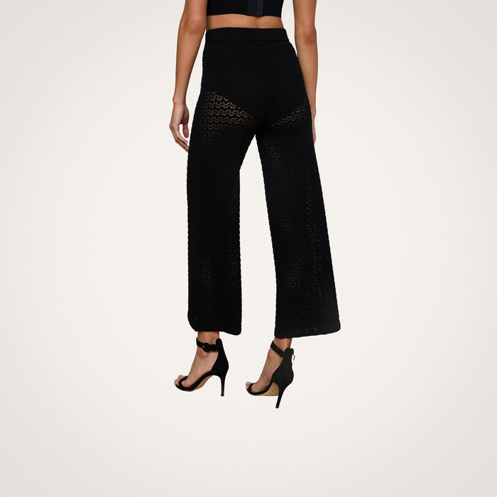 A person wearing Gaelle Pointelle Knit Pants in black, featuring a wide-leg silhouette and drawstring waist, paired with black high-heeled sandals and shown from the back against a plain light background.