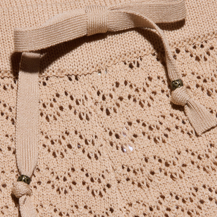 Close-up of a lightweight beige pointelle knit fabric featuring a decorative bow with metallic bead accents at the ends. The lacy hole pattern and delicate details make it perfect for vacation style or pairing with the Gaelle Pointelle Knit Pant.