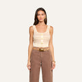 A woman wears the Galene Tank, a sleeveless beige cropped knit top with three decorative buttons, paired with brown high-waisted pants, a wide belt with a brass buckle, gold earrings, and a bracelet, as she stands against an off-white background.