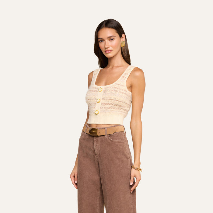 A woman wearing the Galene Tank—a cropped knit top with eyelash texture, three decorative buttons, and wide straps—paired with high-waisted brown corduroy pants and a tan belt, poses against a plain background.
