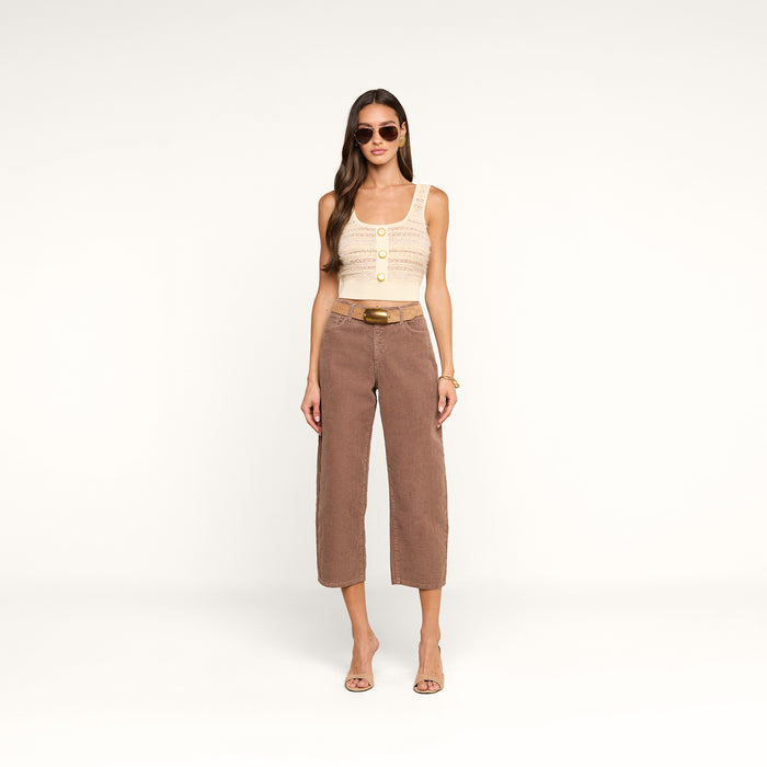 A woman in sunglasses wears the Galene Tank, a sleeveless beige top with decorative buttons, paired with brown belted cropped pants and heeled sandals, standing against a plain white background.