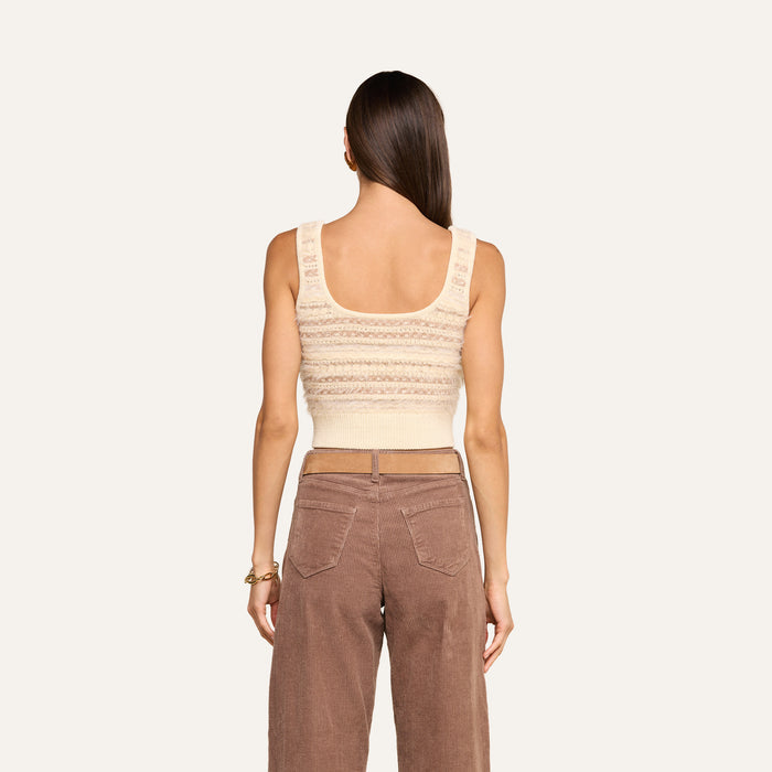 A woman with long brown hair is seen from behind, wearing the cream cropped Galene Tank featuring decorative buttons and high-waisted brown pants, standing against a plain light background.
