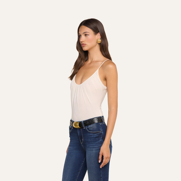 Woman with long brown hair stands in profile, wearing the Gelina Mesh Bodysuit—a sleeveless beige bodysuit—paired with blue jeans featuring a black belt and gold buckle, and accessorized with round gold earrings, against a plain light background.