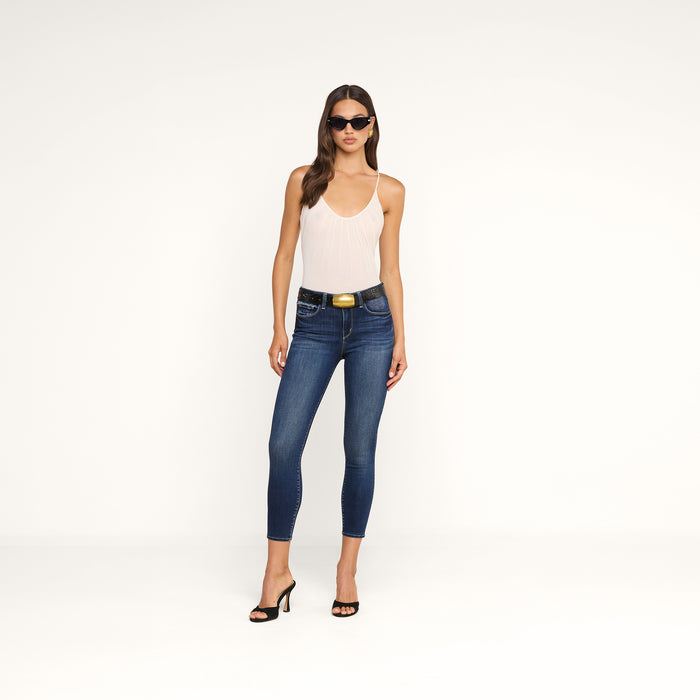 A woman wearing sunglasses, the Gelina Mesh Bodysuit—a light-colored sleeveless piece with adjustable straps—paired with dark blue skinny jeans featuring a belt and black heeled sandals, stands against a plain white background.
