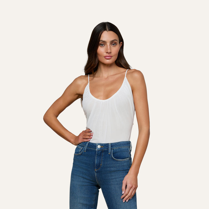 A woman with long brown hair wears the Gelina Ruched Bodysuit, featuring a white mesh design and adjustable straps, paired with blue jeans as she poses with one hand on her hip against a plain light background.