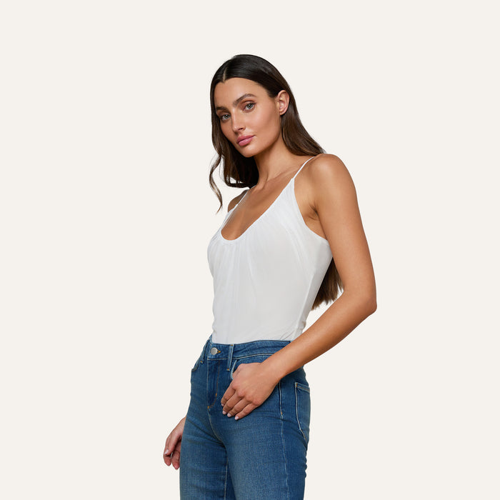 A woman with long dark hair stands confidently against a plain light background, wearing the Gelina Ruched Bodysuit with adjustable straps and blue jeans. She poses in a relaxed manner, looking directly at the camera.