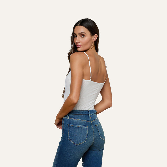 A woman with long brown hair wears the Gelina Ruched Bodysuit, a white bodysuit with adjustable straps, paired with blue jeans. She stands slightly turned to the side, looking over her shoulder against a plain light background.