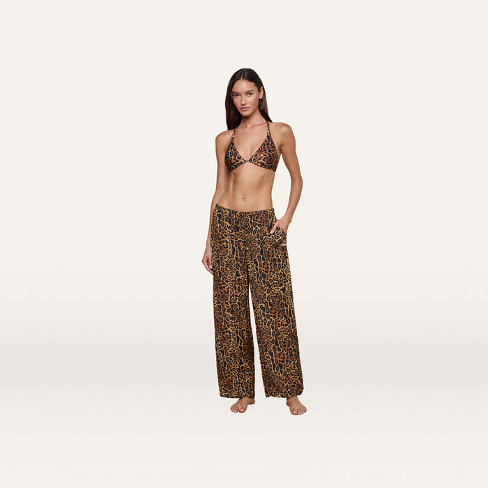 A woman stands barefoot, posing against a plain white background in the Geraldine Cover-Up Pant—wide-leg, pull-on fit pants featuring a matching leopard print bikini top—perfect for resort wear.