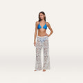 A woman stands barefoot against a plain light background, wearing a blue bikini top paired with the Geraldine Cover-Up Pant—white bohemian pants featuring a paisley crochet pattern.