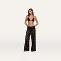 A woman stands with her hands in her pockets against a plain white background, wearing a black bikini top and the Geraldine Cover-Up Pant—a pair of sheer black lace bohemian pants.