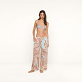 A woman stands barefoot in resort wear, featuring a paisley print bikini top paired with the Geraldine Cover-Up Pant—loose and relaxed in silhouette, showcasing light blue and brown tones—against a plain white background.