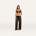 A woman stands barefoot in sheer black Geraldine Cover-Up Pants and a black bikini top, evoking a chic bohemian vibe against a plain white background.