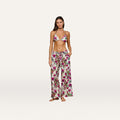 A woman stands barefoot against a plain light background, her hands tucked in her pockets, wearing the Geraldine Cover-Up Pant—a pair of loose linen resort pants with a tie waist in a rose leopard print—paired with a matching floral bikini top.