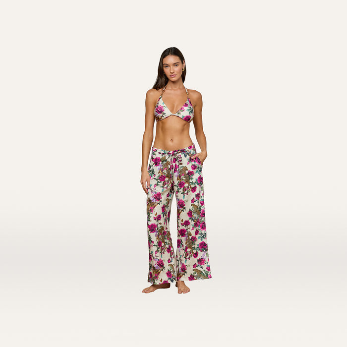 A woman stands barefoot against a plain light background, her hands tucked in her pockets, wearing the Geraldine Cover-Up Pant—a pair of loose linen resort pants with a tie waist in a rose leopard print—paired with a matching floral bikini top.