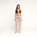 A woman stands against a plain background, facing forward with a neutral expression, wearing a black bikini top paired with the Geraldine Cover-Up Pant—white sheer lace bohemian pants.