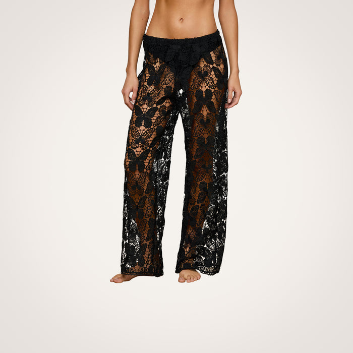 A person stands barefoot against a plain white background, wearing the Geraldine Cover-Up Pant. These black, see-through butterfly crochet lace pants feature a brown underlayer, a loose wide-leg fit, and a low waist—perfect for effortless bohemian resort wear style.