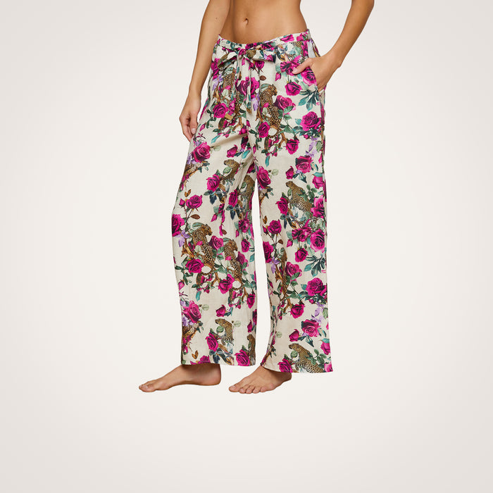 A person stands barefoot against a plain white background, wearing the Geraldine Cover-Up Pant—wide-leg linen resort pants featuring pink and purple flowers with green leaves and a comfortable tie waist.
