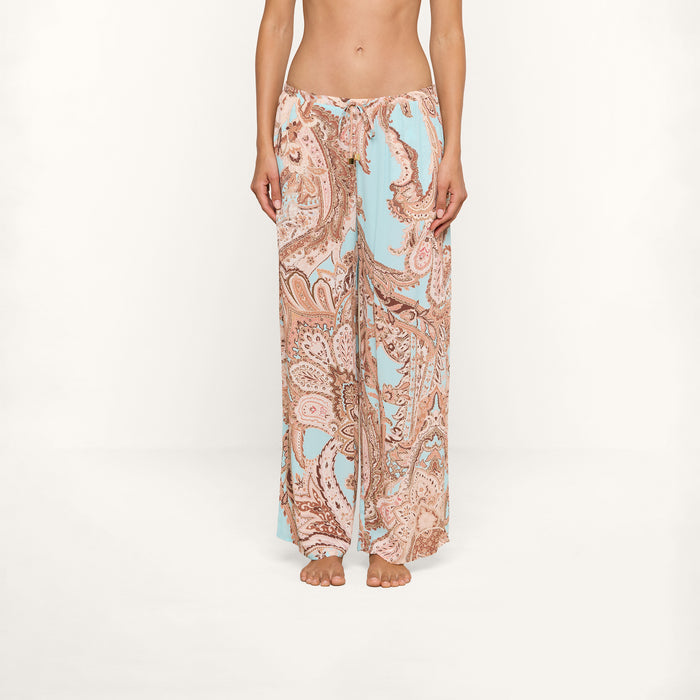 A person stands barefoot wearing the Geraldine Cover-Up Pant, featuring a wide-leg, light blue and tan paisley print with a relaxed silhouette. The image is cropped from the chest down, showcasing the pants against a plain white background.