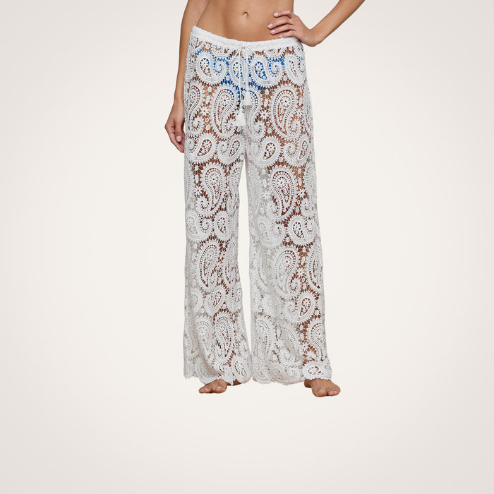 A person is modeling the Geraldine Cover-Up Pant—sheer, white bohemian pants featuring a paisley lace pattern and a loose fit with a drawstring waist, perfect for resort wear. The model is barefoot against a plain background.