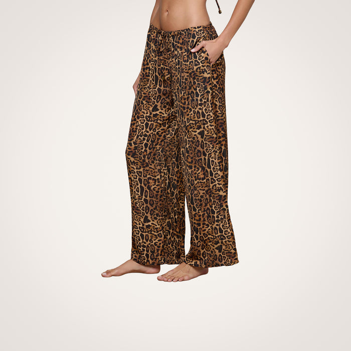 A person stands barefoot against a plain white background, modeling the Geraldine Cover-Up Pant—a loose, resort wear style featuring a wide-leg leopard print design, side pockets, and a comfortable pull-on fit with a drawstring waist. The top half of the body is not visible.