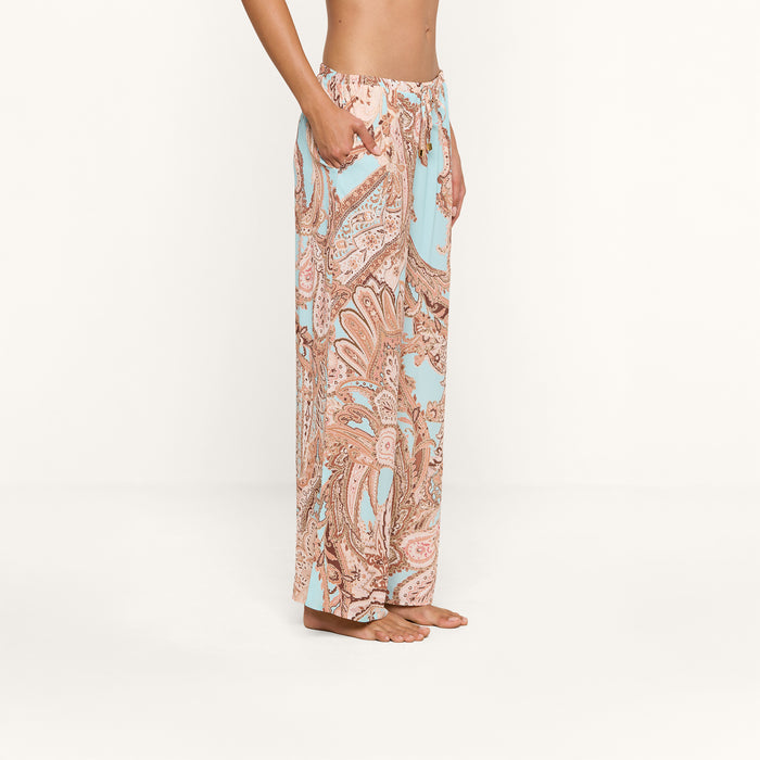 A person stands barefoot in side profile, wearing the Geraldine Cover-Up Pant—a high-waisted, loose-fitting style adorned with a pastel paisley print in pink and light blue hues. With its relaxed silhouette, this stylish piece embodies the effortless elegance of resort wear against a plain white background.