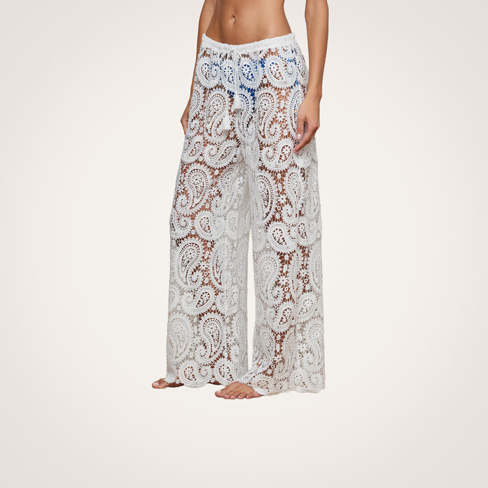 A person stands barefoot against a plain background, wearing the Geraldine Cover-Up Pant. These white, sheer bohemian pants feature an intricate paisley crochet pattern and a loose, see-through design—perfect for resort wear.