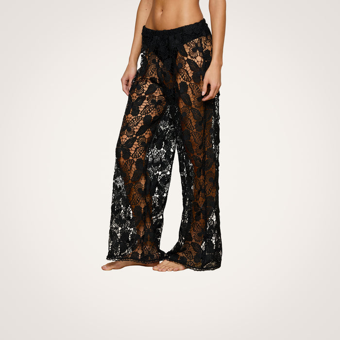 Model wearing the Geraldine Cover-Up Pant—black, sheer, lace wide-leg pants featuring a floral pattern and bohemian flair—standing barefoot against a plain white background.