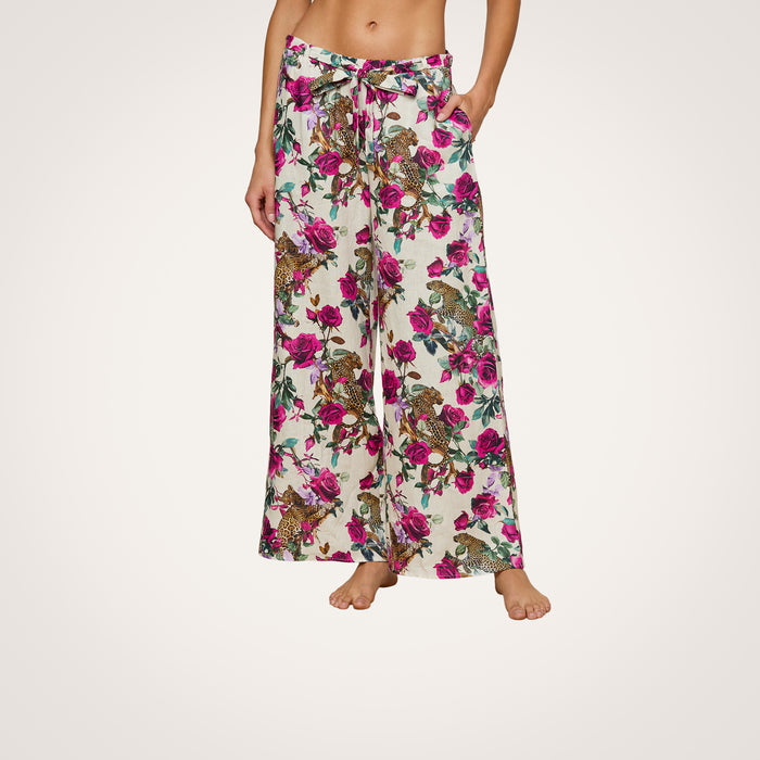 A woman models the Geraldine Cover-Up Pant, a wide-leg, high-waisted linen resort style featuring a pink and green rose leopard print. The pants include a tie waist and side pockets. She stands barefoot against a plain background.