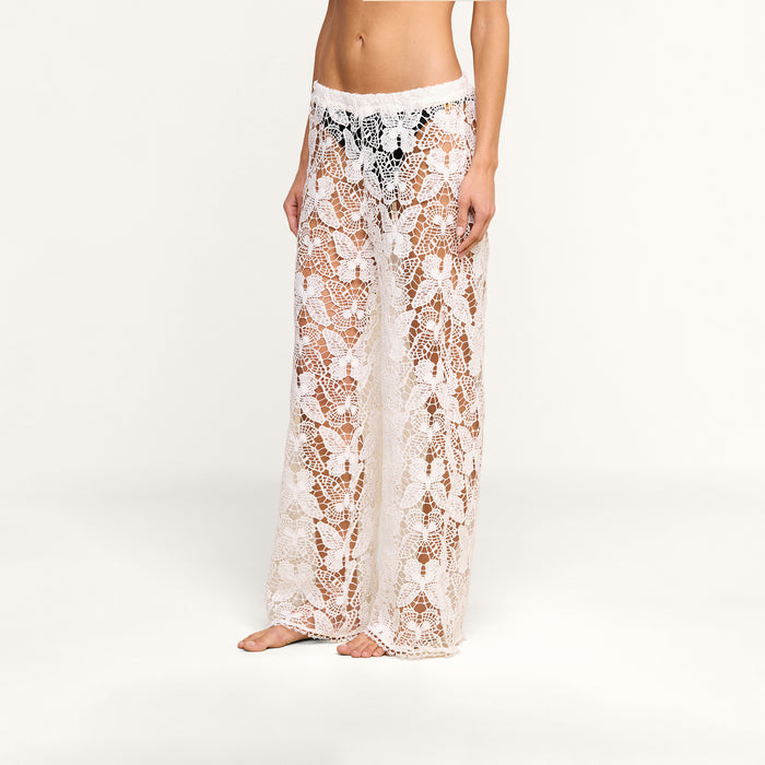 A person wearing the Geraldine Cover-Up Pant, featuring a sheer white bohemian crochet lace design with a floral pattern, stands barefoot against a plain white background.