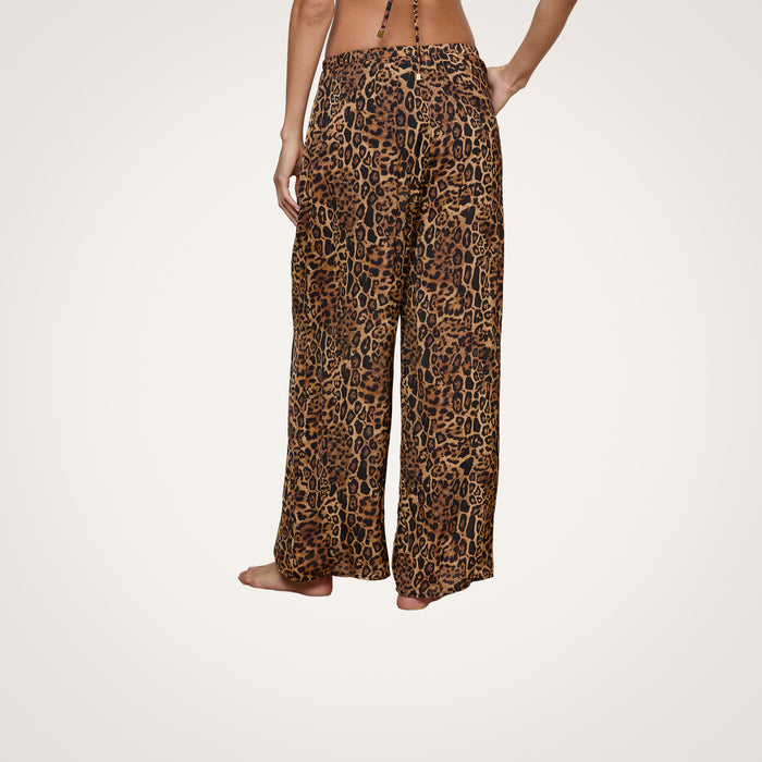 A person stands barefoot, viewed from the back, wearing the Geraldine Cover-Up Pant—a wide-leg, leopard print style with a relaxed pull-on fit. The upper body is mostly out of frame.