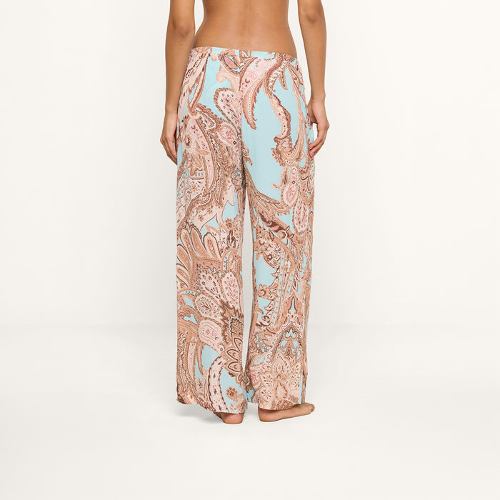 A person stands barefoot, facing away, wearing the Geraldine Cover-Up Pant—a loose-fitting, wide-leg style featuring a light blue and tan paisley print. The relaxed silhouette of the pants adds to the effortless feel against the plain white background.