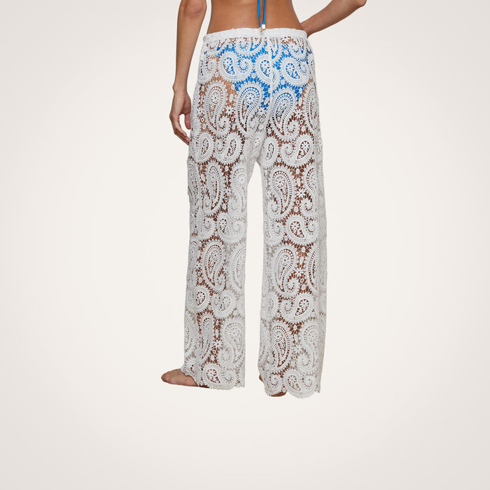 A person stands barefoot against a plain background, wearing the Geraldine Cover-Up Pant—a white, sheer, paisley crochet bohemian style—paired with a blue bikini top.