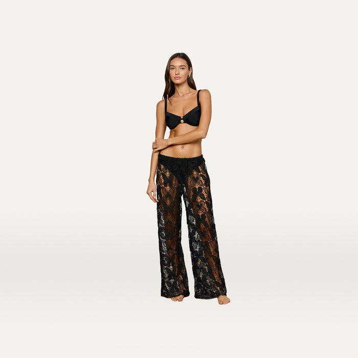 A woman stands barefoot wearing a black bra and the Geraldine Cover-Up Pant—sheer black lace with a nude underlay—embodying effortless bohemian style as she poses against a plain white background.