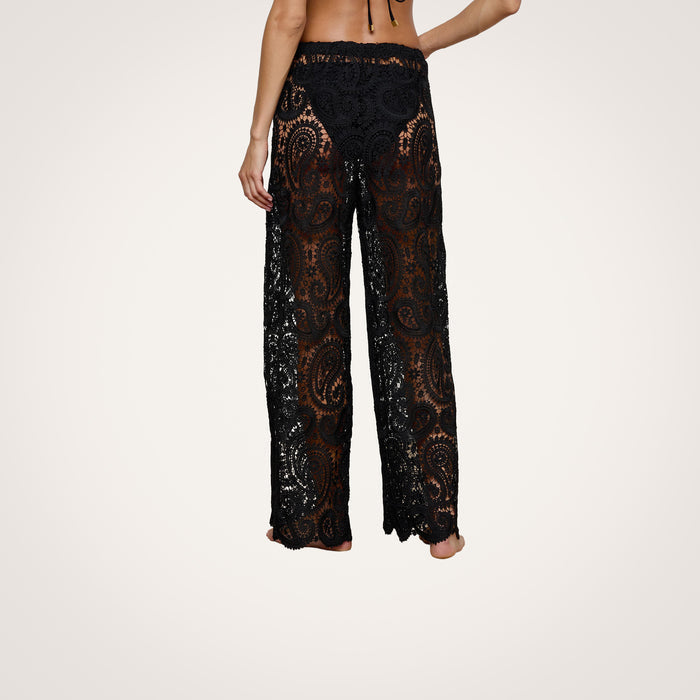 A person stands barefoot, seen from behind, wearing the Geraldine Cover-Up Pant—bohemian-style pants crafted from black lace with a paisley crochet pattern. The upper body is mostly out of frame against a plain, light-colored background.